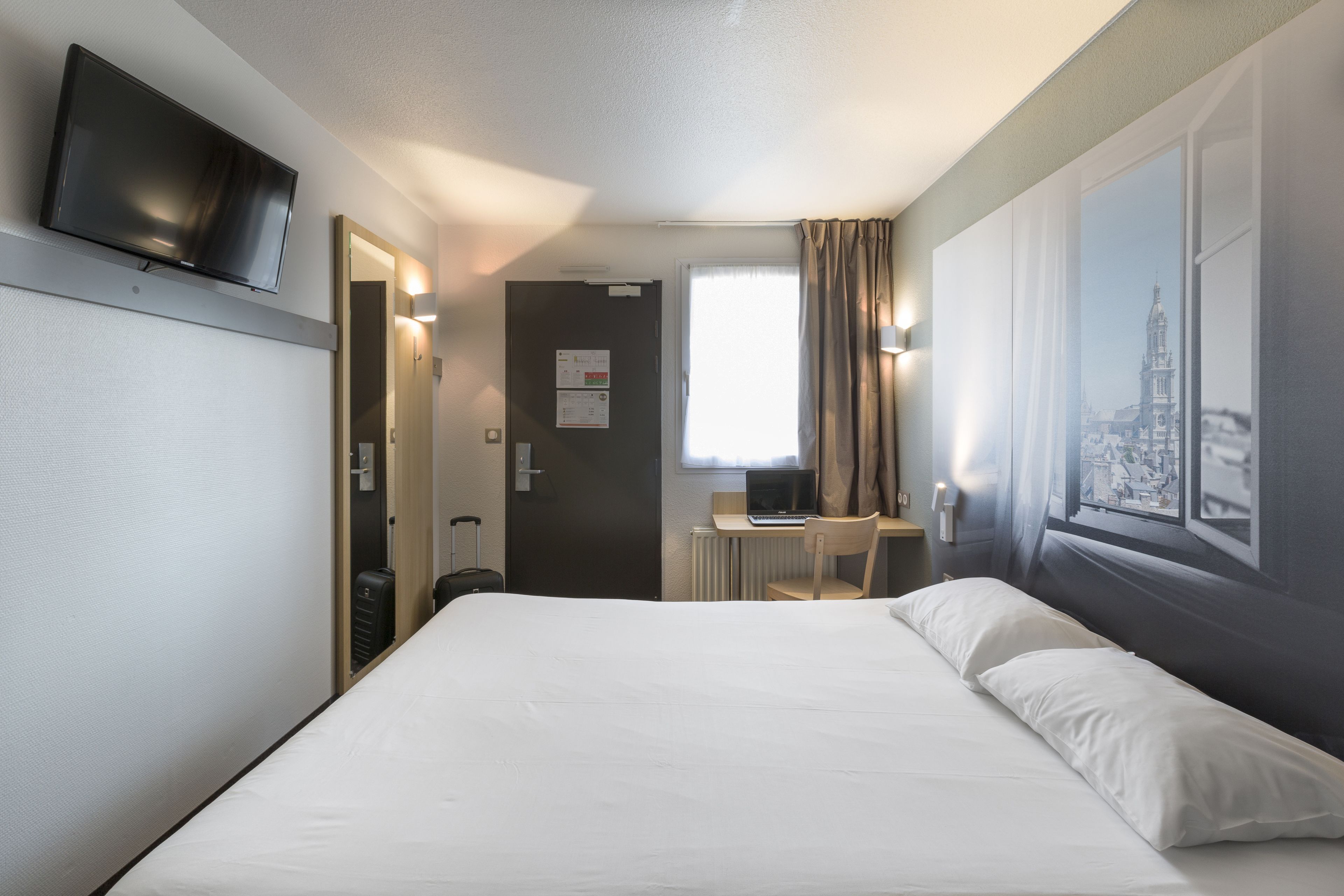 A hotel room with a bed, a desk with a laptop, a television, and a window with curtains.