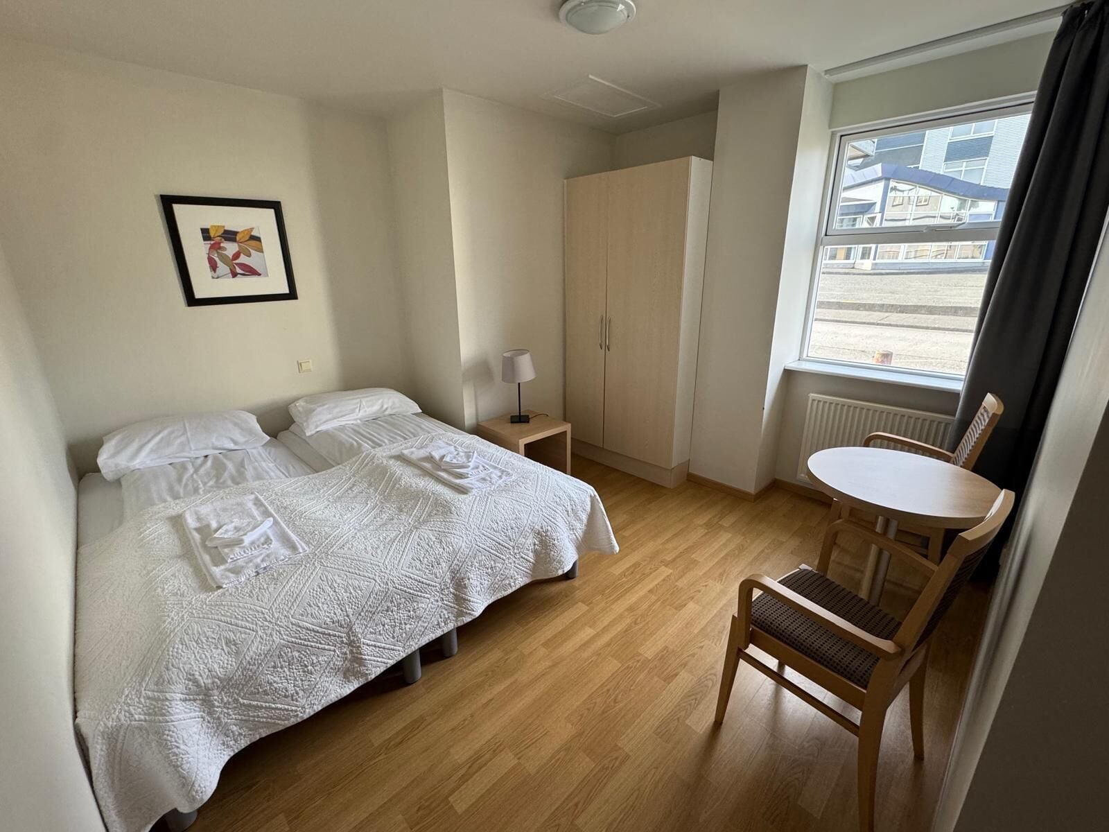 A hotel room with a bed, a chair, a small table, a wardrobe, and a window with a view of buildings.
