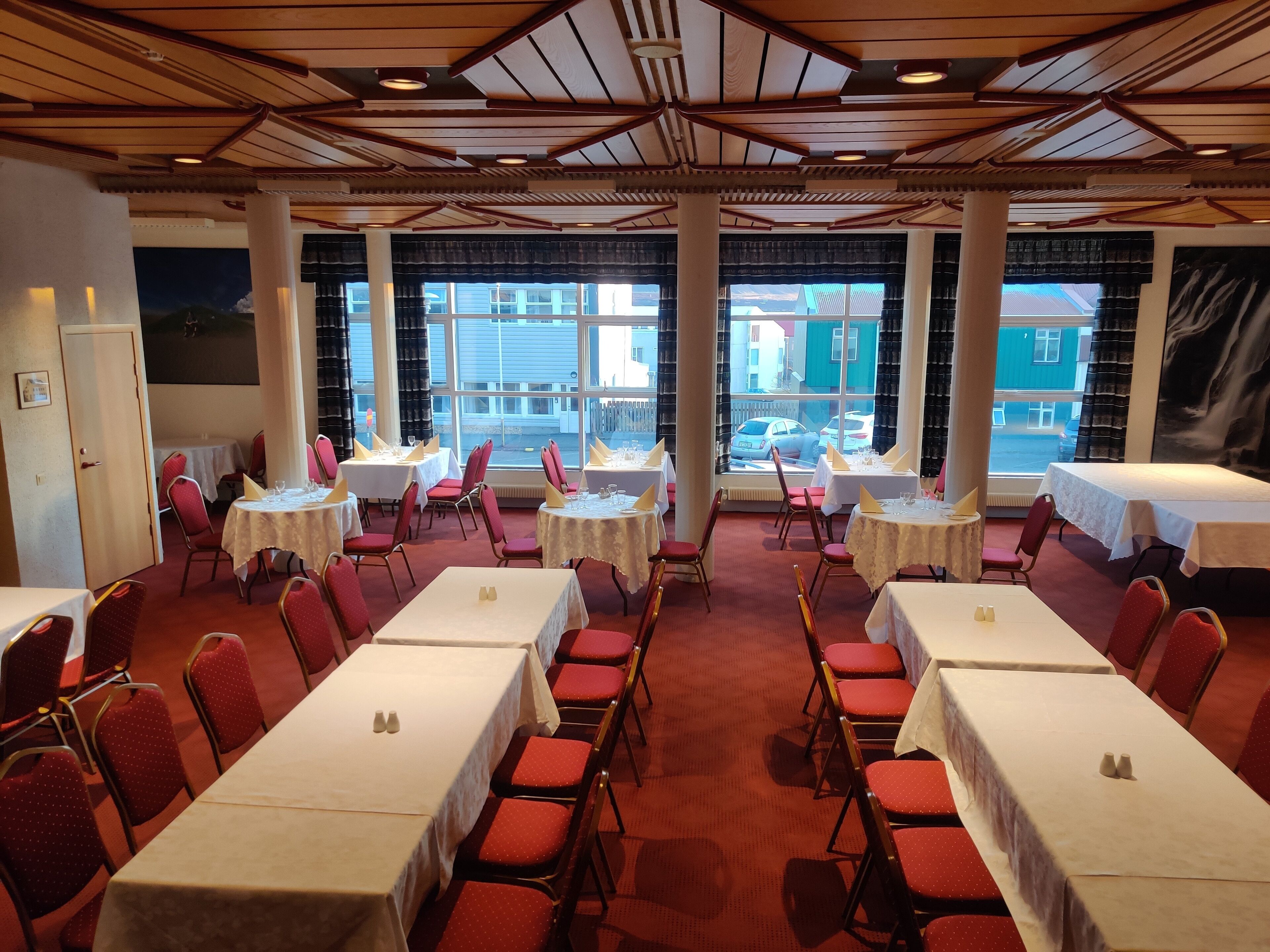 Un salón de conferencias con varias mesas y sillas, amplias ventanas que ofrecen vistas a edificios y automóviles, y un techo de madera con vigas a la vista.