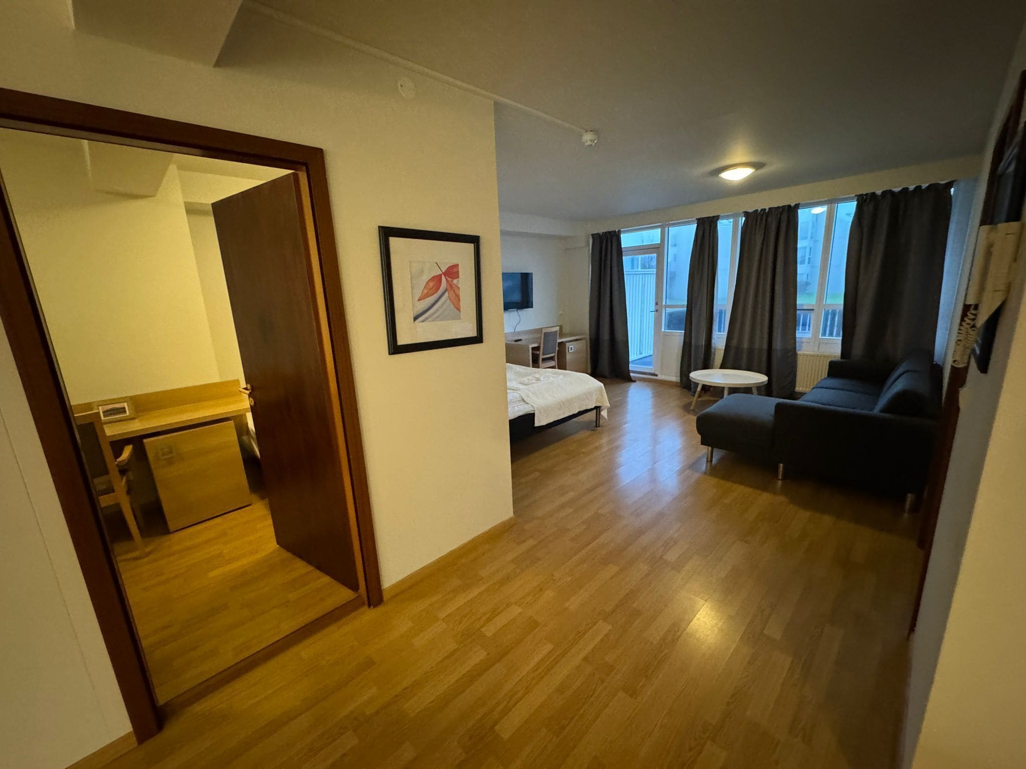 A hotel room with a wooden floor, a bed, a desk, a chair, a sofa, and a TV.