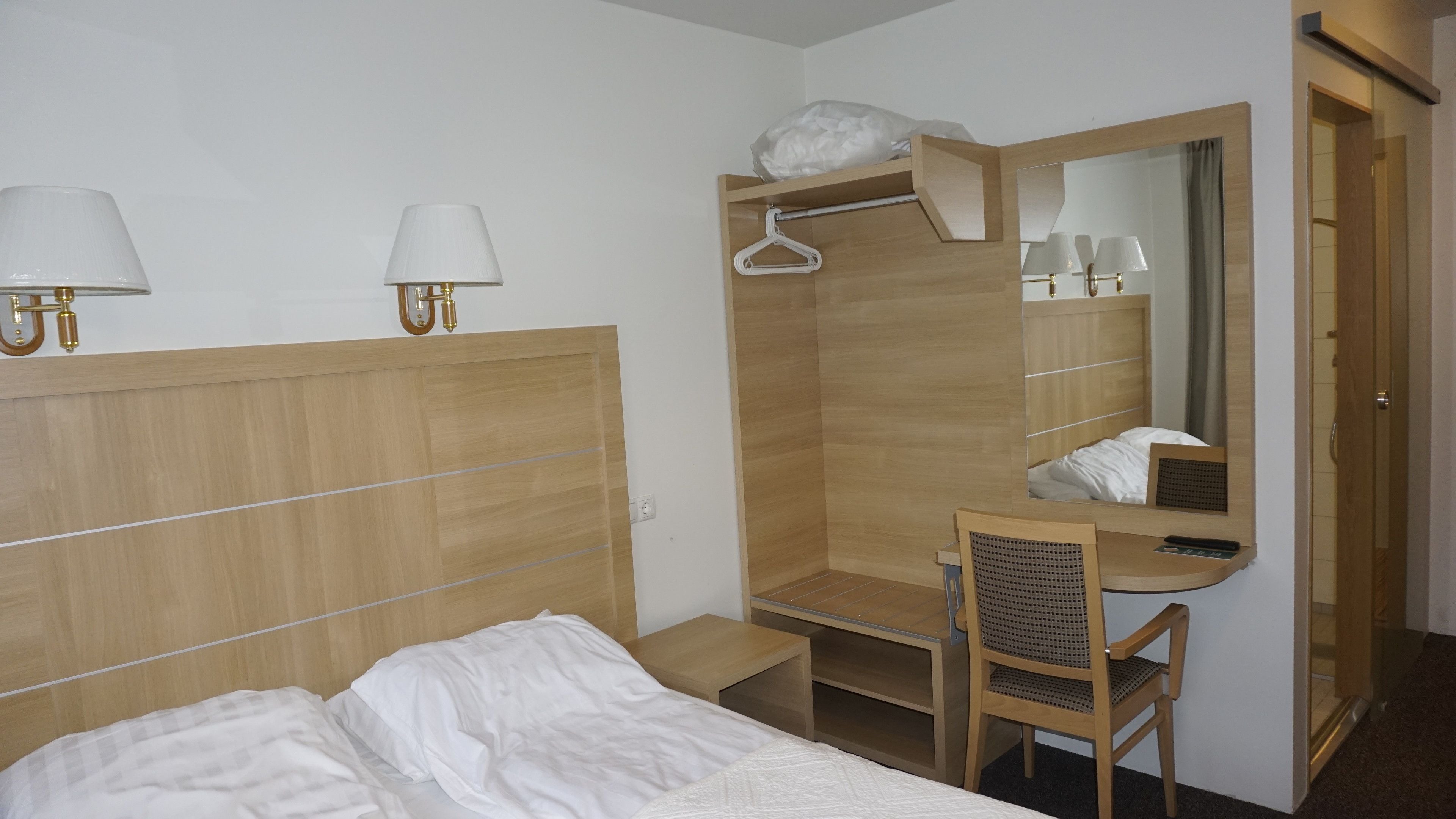 A hotel room with a wooden bed, a desk with a chair, a mirror, and two wall-mounted lamps.