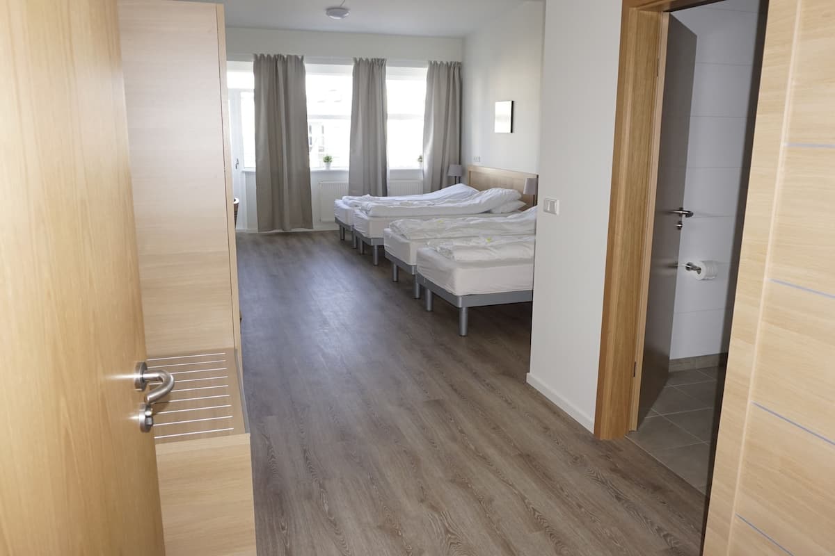 A hotel room with a wooden floor, a bed, a window with curtains, and a door.