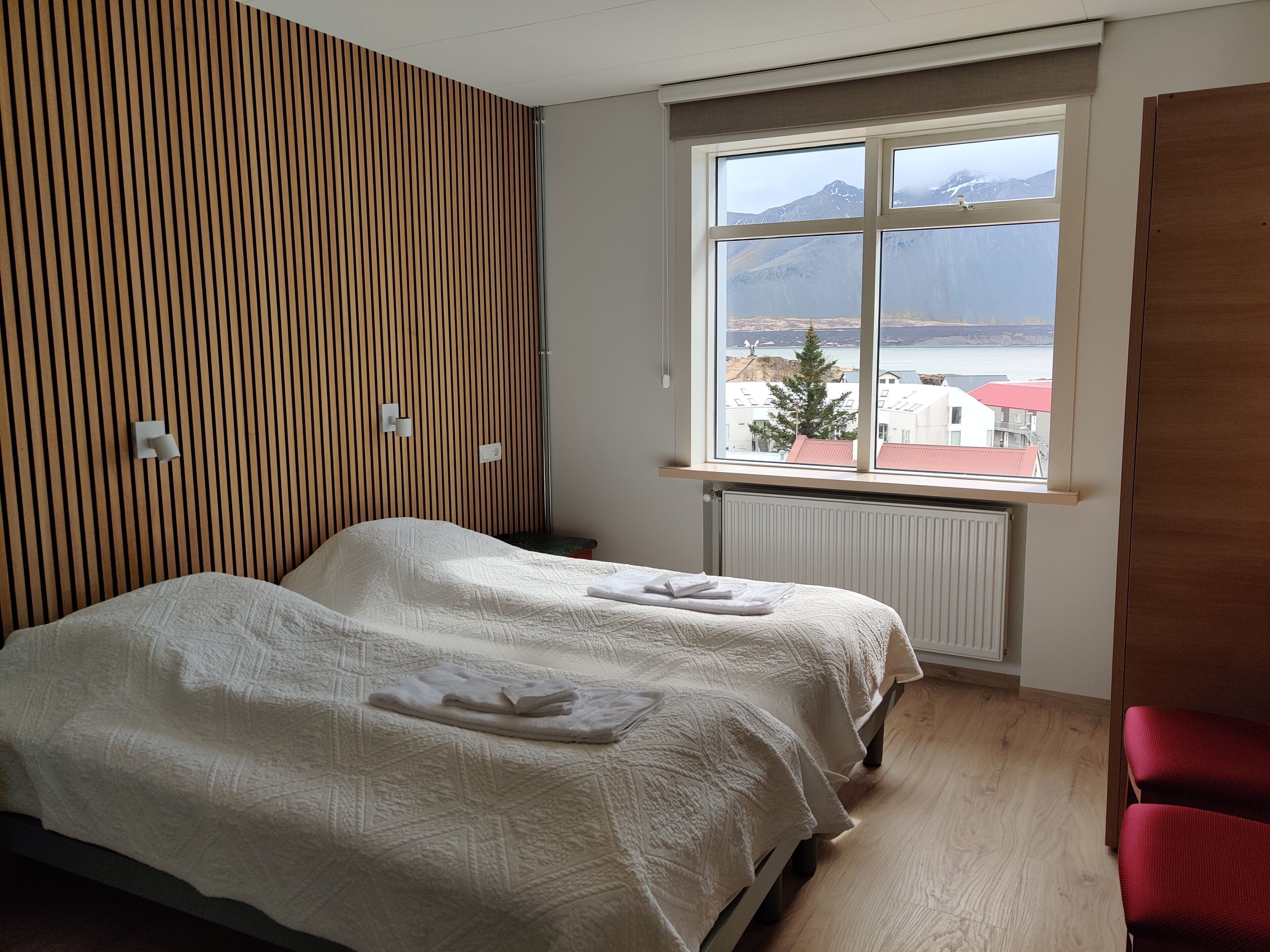 A hotel room with two beds, a wooden slat wall, a large window with a mountain view, and a red chair.
