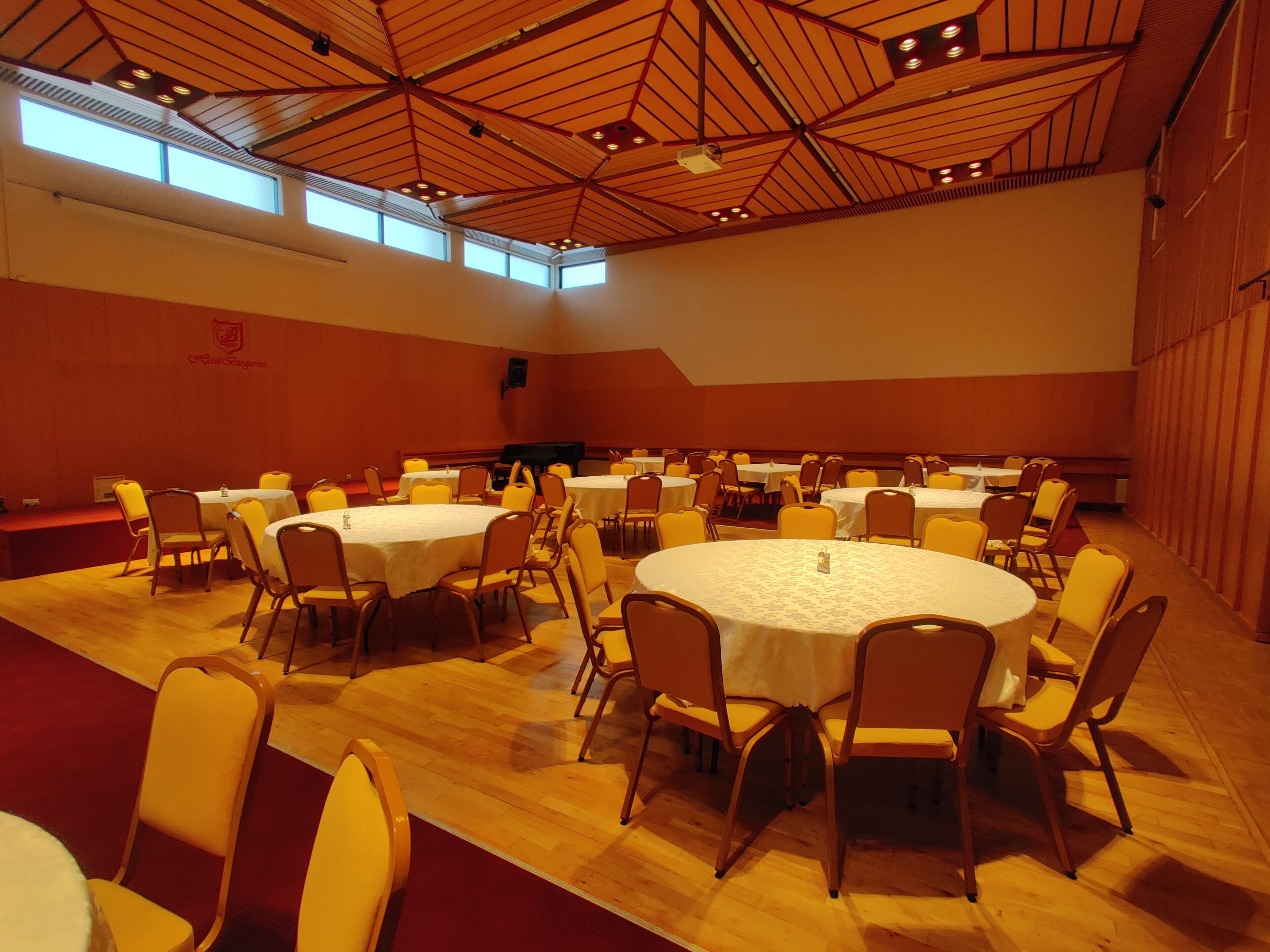 Un amplio salón de conferencias con varias mesas redondas y sillas, techos altos con vigas de madera y ventanas grandes que dejan entrar luz natural.