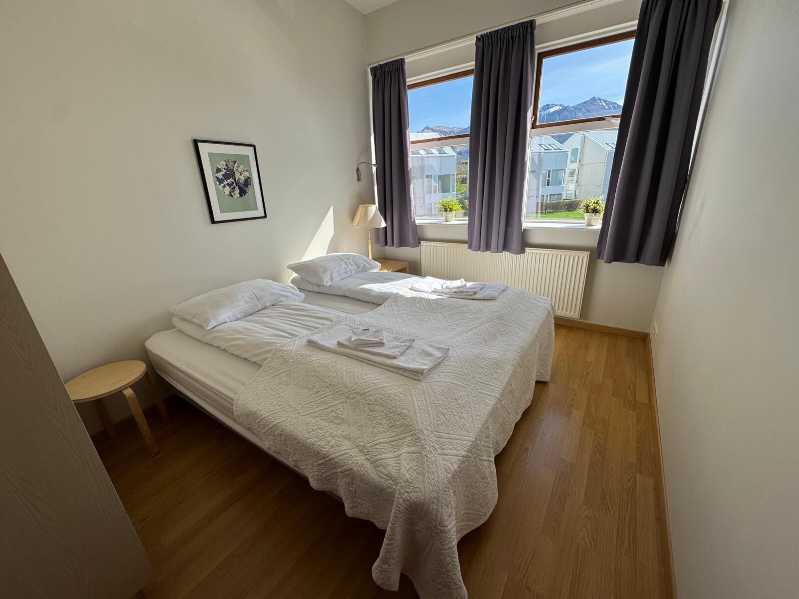 A hotel room with two beds, a small table, a framed picture on the wall, and a view of buildings and mountains through the window.