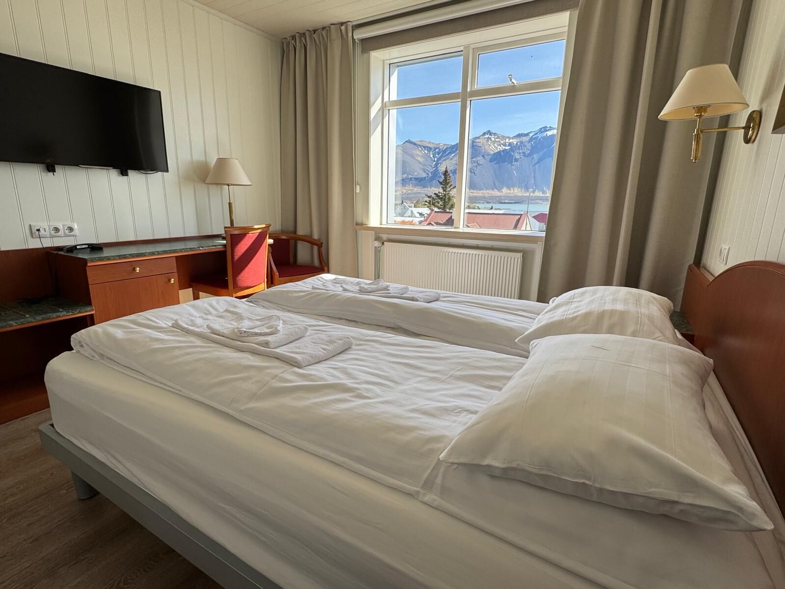 Habitación de hotel con dos camas, un escritorio, una silla, un televisor y ventana con vista a la montaña.