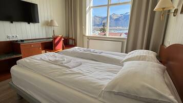 Habitación de hotel con dos camas, un escritorio, una silla, un televisor y ventana con vista a la montaña.