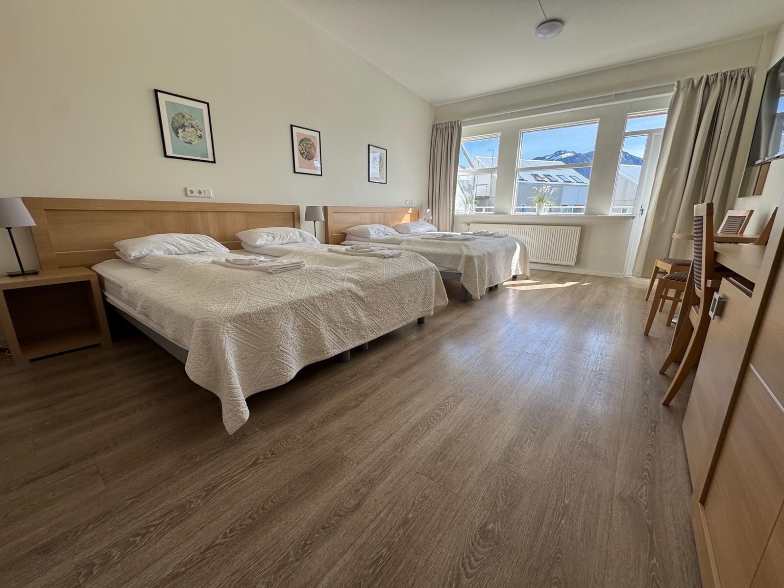 A hotel room with two beds, a wooden headboard, a nightstand, a desk, and a view of mountains through large windows.