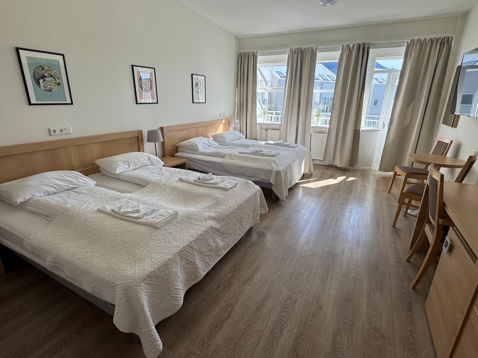 A hotel room with two beds, a wooden floor, a dining area with a table and chairs, and large windows with curtains.