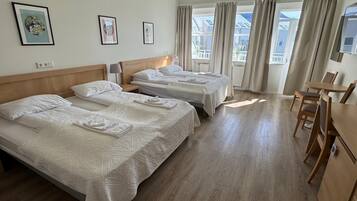 A hotel room with two beds, a wooden floor, a dining area with a table and chairs, and large windows with curtains.