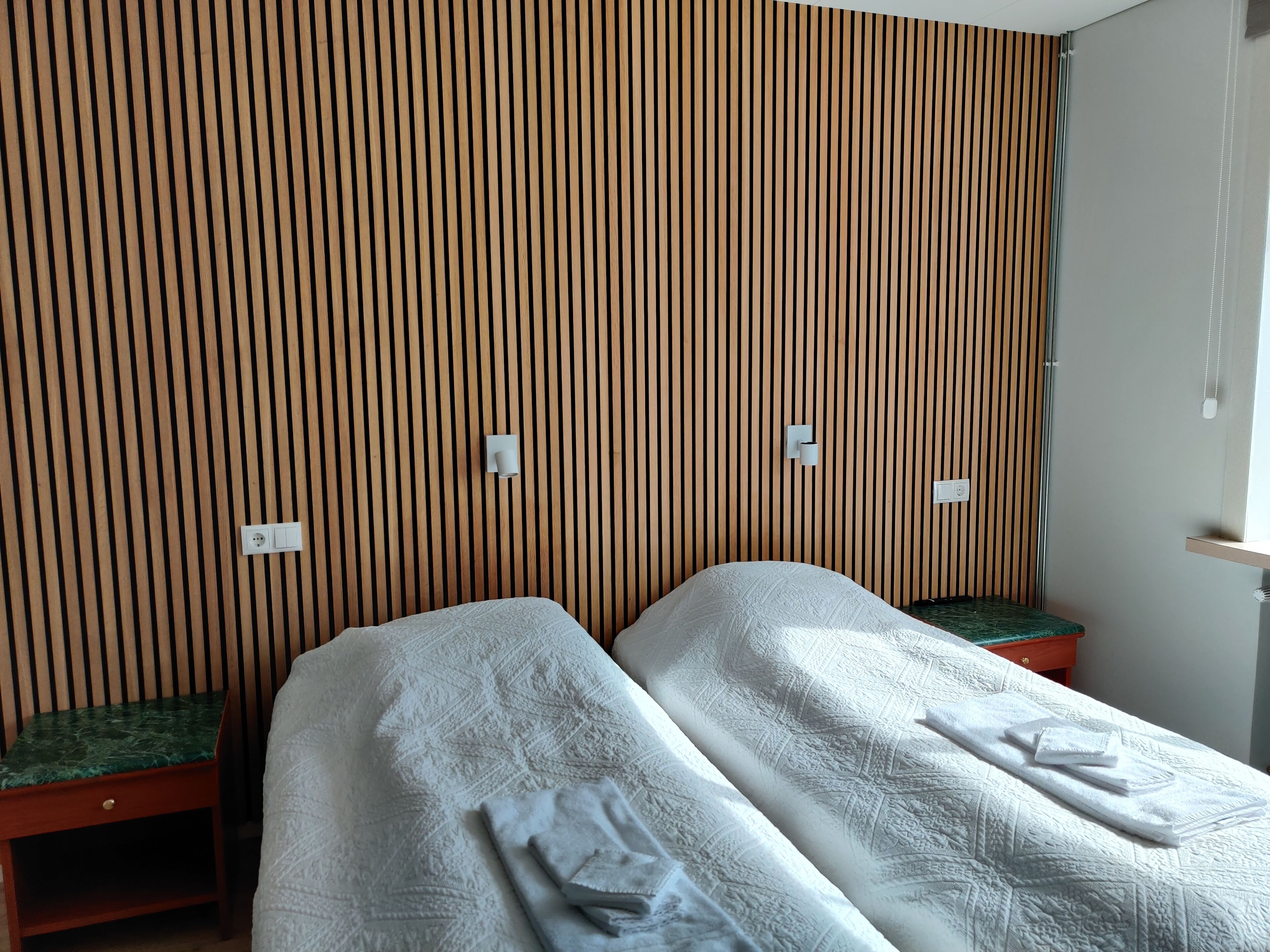 Quarto com duas camas, paredes de ripas de madeira e uma pequena mesa com almofada verde.