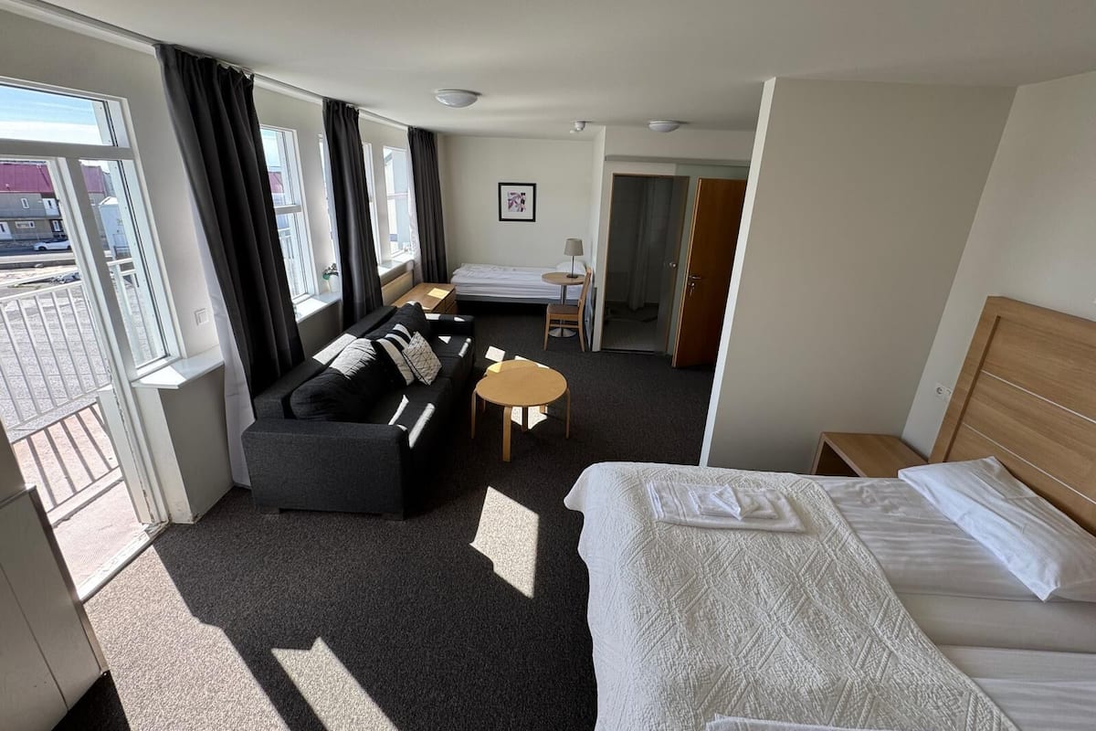 A hotel room with a bed, a sofa, a small table, and a desk. There is a window with curtains, a door, and a picture on the wall.