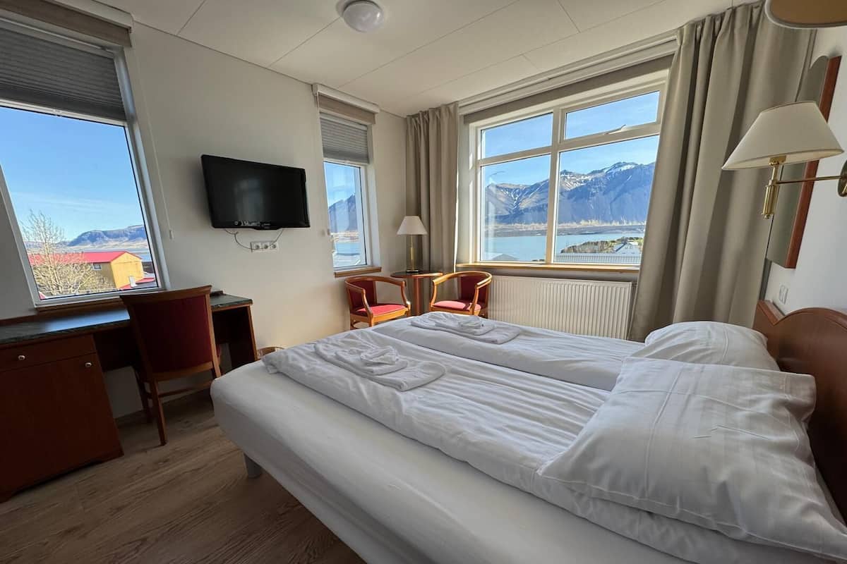 A hotel room with a large bed, a desk, a chair, a TV, and a view of mountains and a lake.