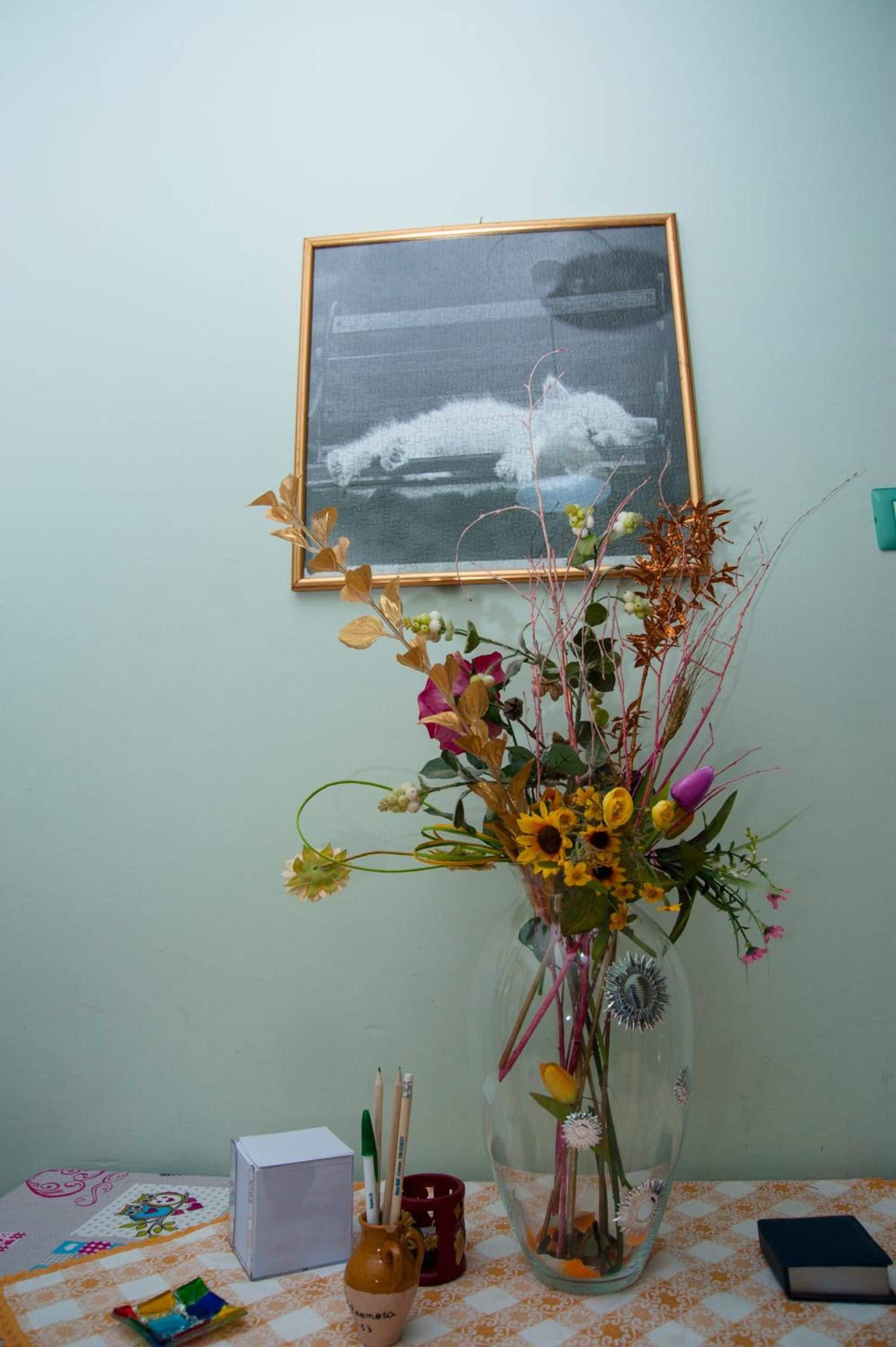 A room with a table, a vase with flowers, a framed picture on the wall, and a few items on the table.