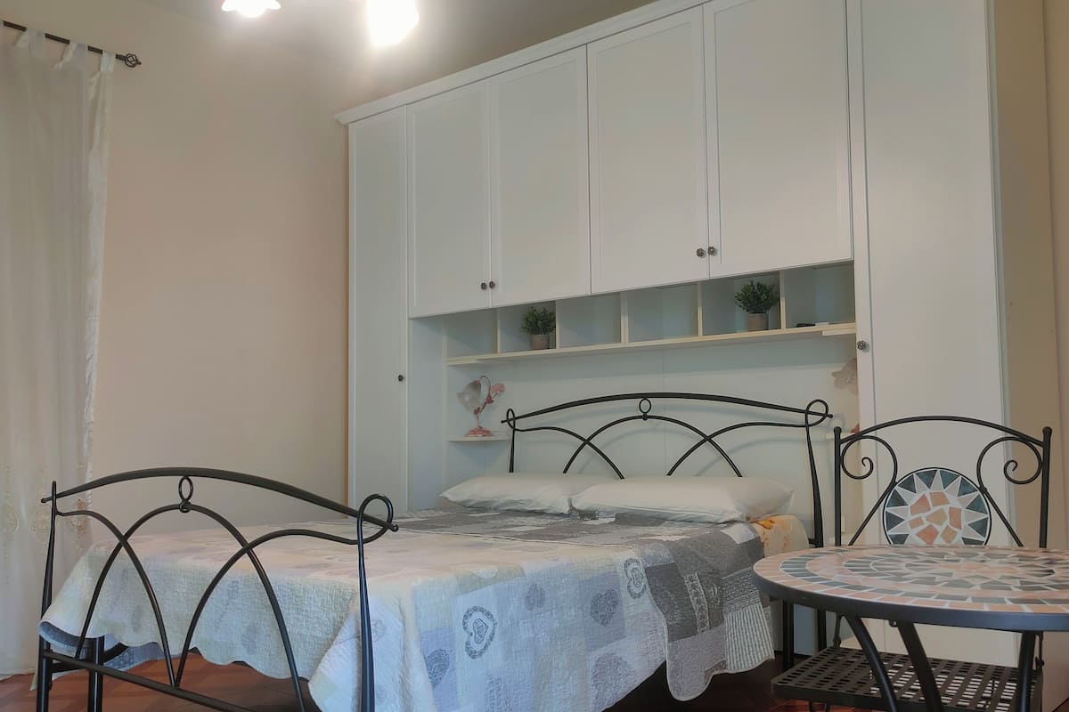 Un dormitorio con una cama, dos mesitas de noche, una mesita redonda pequeña y un armario con estantes.