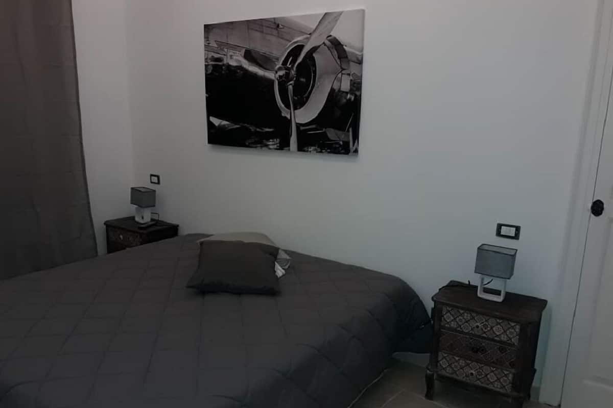 A bedroom with a large bed, a nightstand with a lamp, a small table with a lamp, and a wall-mounted picture.
