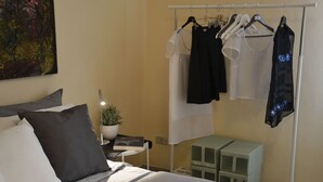 A hotel room with a bed, a bedside table with a book and a plant, a clothing rack with various garments, and a small green cabinet. - Altera Domus (Naples)