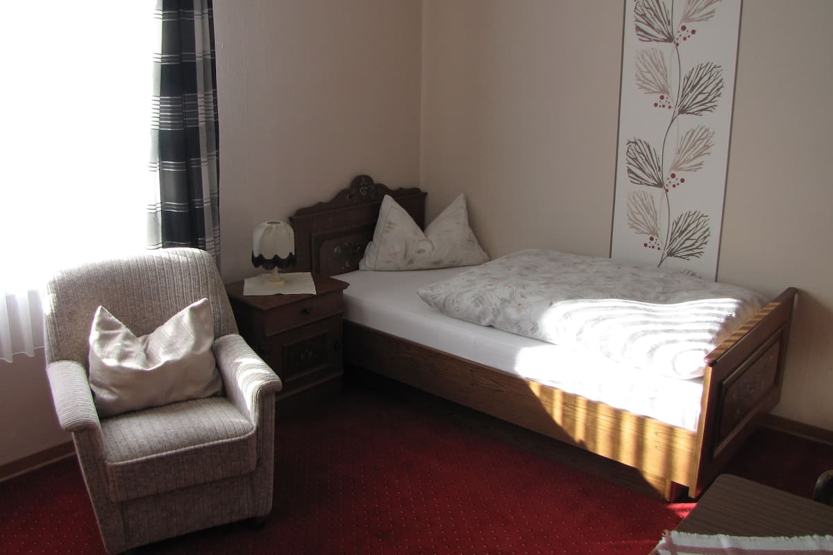 A small bedroom with a single bed, a chair, a nightstand, and a window with sheer curtains.
