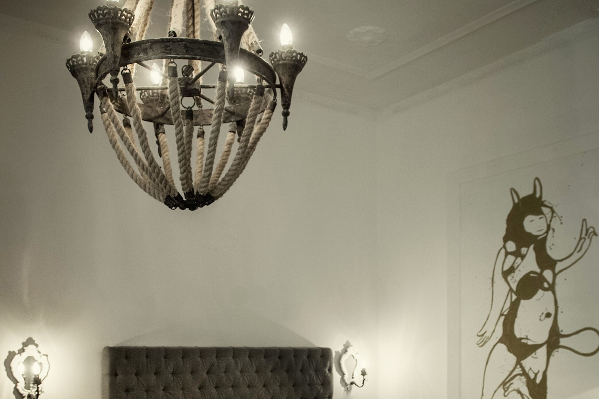 A bedroom with a tufted headboard, a white bedspread, a wooden nightstand with a lamp, a wooden side table, and a chandelier.