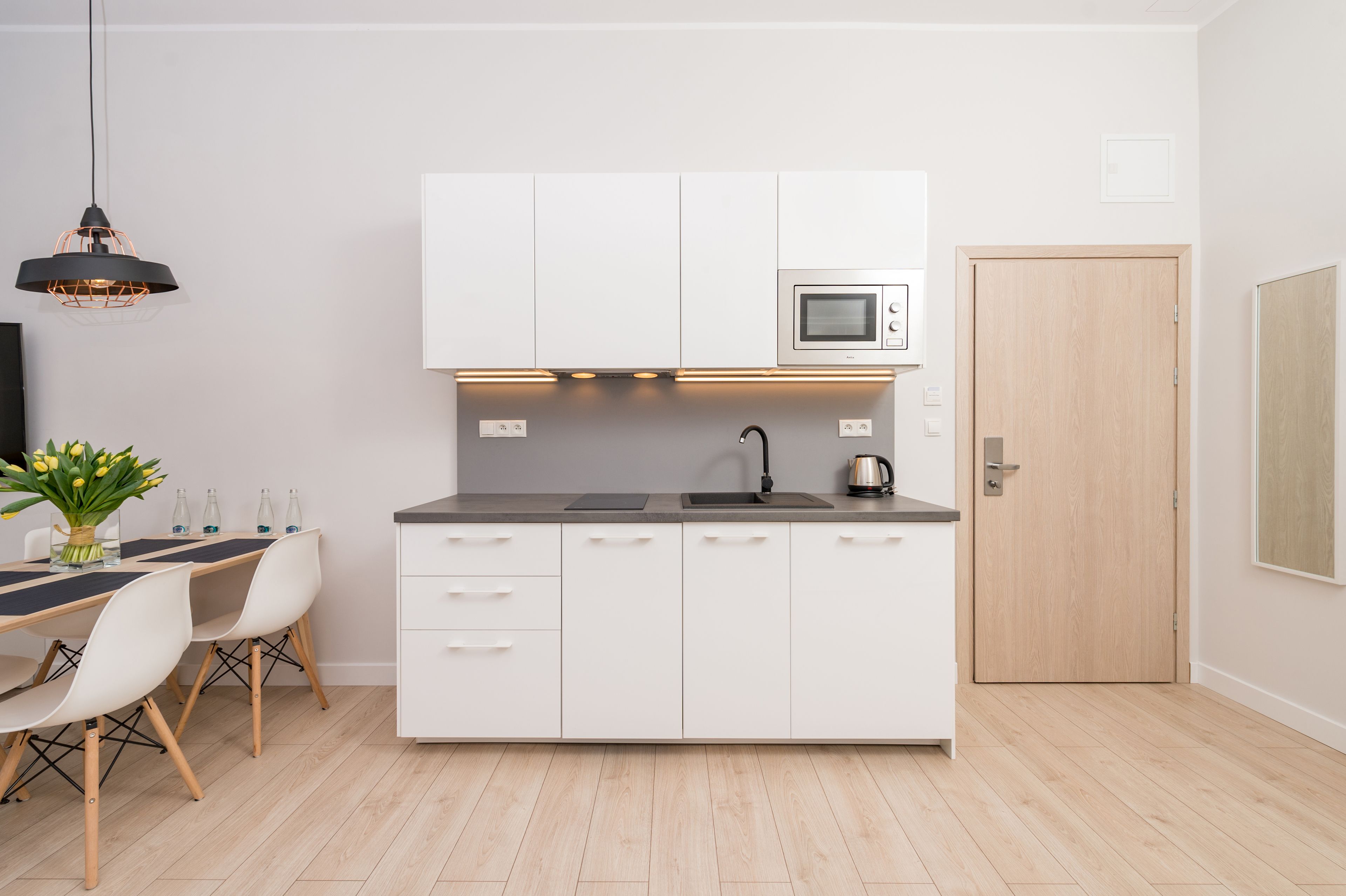 A compact kitchen with white cabinets, a microwave, a kettle, and a dining area with a table and chairs.