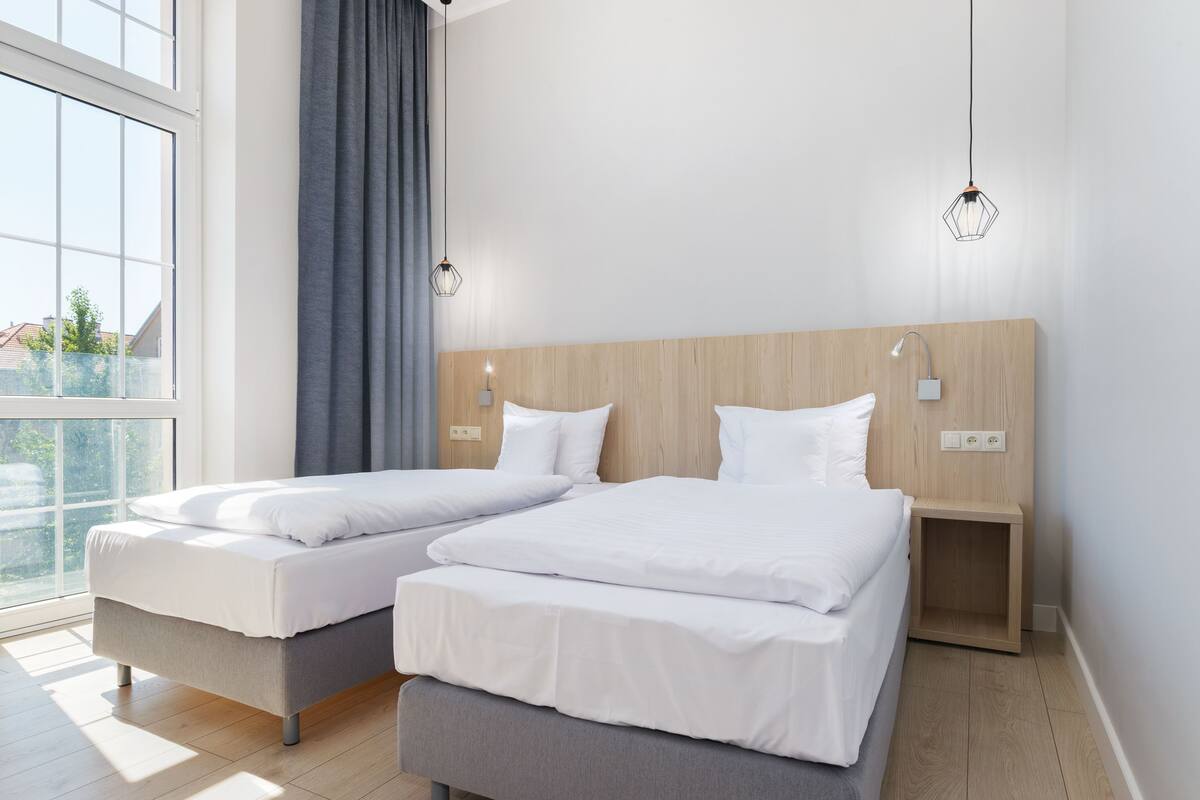 Une chambre moderne avec deux lits simples, une tête de lit en bois, des lampes de chevet et une grande fenêtre avec des rideaux.
