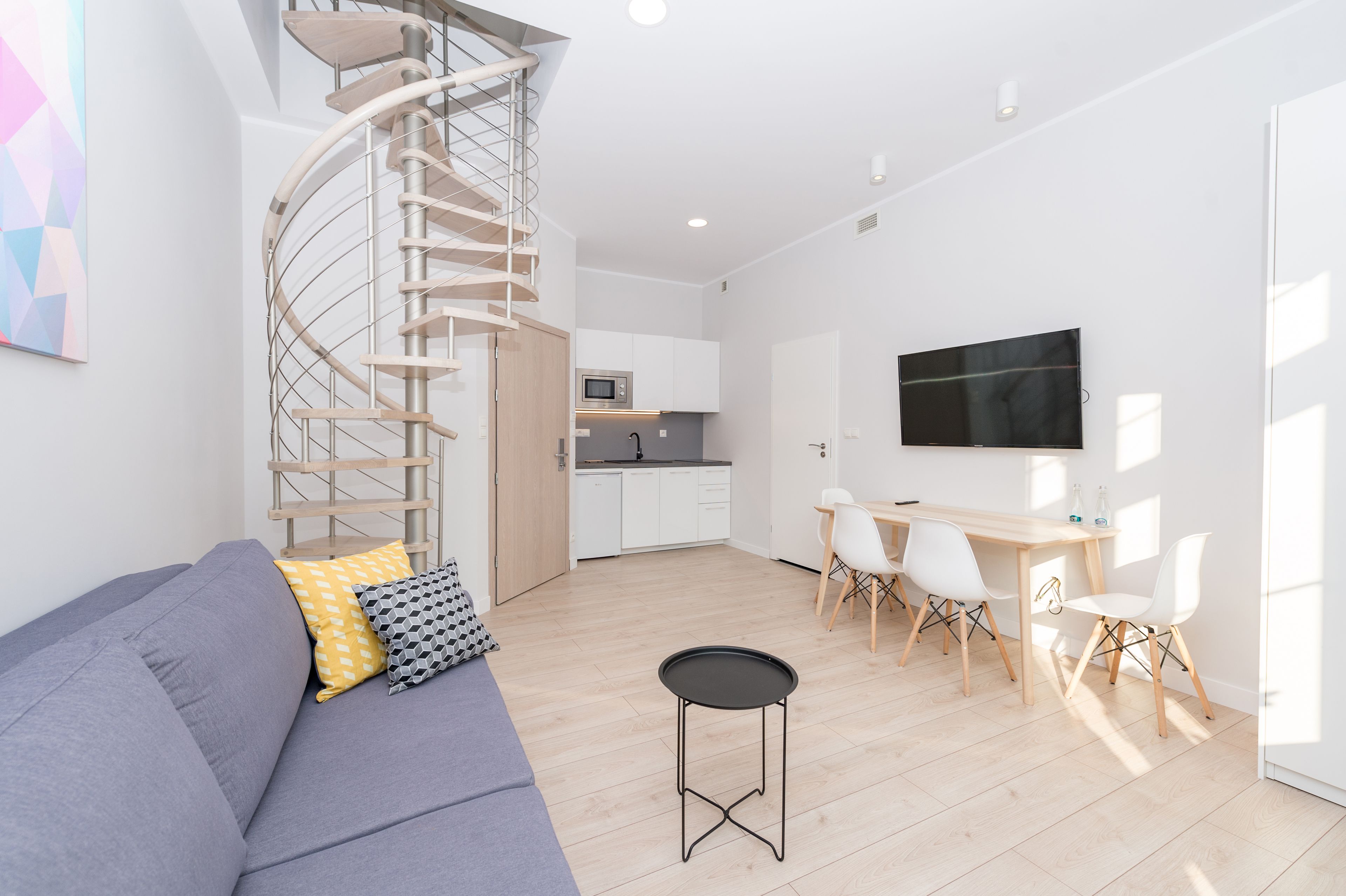 A modern living room with a spiral staircase, a grey sofa, a dining area with a table and chairs, and a kitchen in the background.