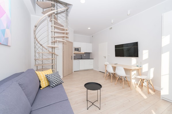 Una sala de estar moderna con una escalera espiral, un sofá gris, una zona de comedor con mesa y sillas, y una cocina al fondo.