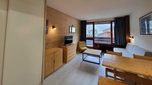 A compact living space with a wooden wall, a television, a sofa, a dining table, and a window offering a view of buildings and mountains. - Southwest studio in Tignes, near slopes, 27 m² (Tignes)