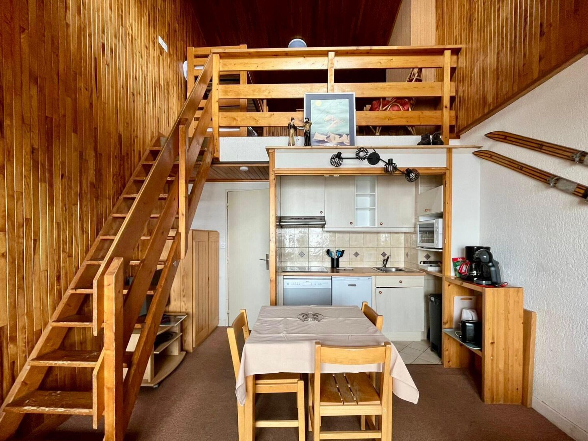 Une pièce compacte à plusieurs niveaux, dotée d’un escalier en bois, d’un coin cuisine, d’un espace repas et d’un rangement mural pour le matériel de ski.