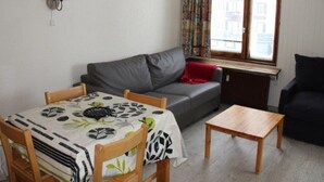 Ein Wohnzimmer mit grauem Sofa, einem Holztisch und einem Esstisch mit einem Tischläufer mit Blätermuster.