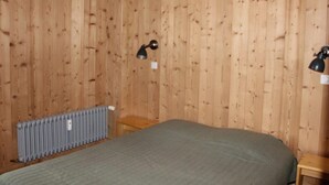 Une pièce aux murs recouverts de lambris en bois, un lit avec une couverture verte, un radiateur et deux lampes fixées au mur.
