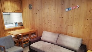 Une pièce chaleureuse avec un mur en bois, un canapé, un fauteuil, une petite table et un coin cuisine visible à travers une fenêtre.