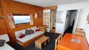 A cozy room with a wooden wall, a sofa with patterned cushions, a small table with an orange tablecloth, and a window with curtains. - 2-room south-facing apartment with wifi 500m from slopes (Tignes)