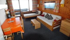 A cozy room with a wooden wall, a sofa, a dining table with chairs, and a window with curtains. - 2-room south-facing apartment with wifi 500m from slopes (Tignes)