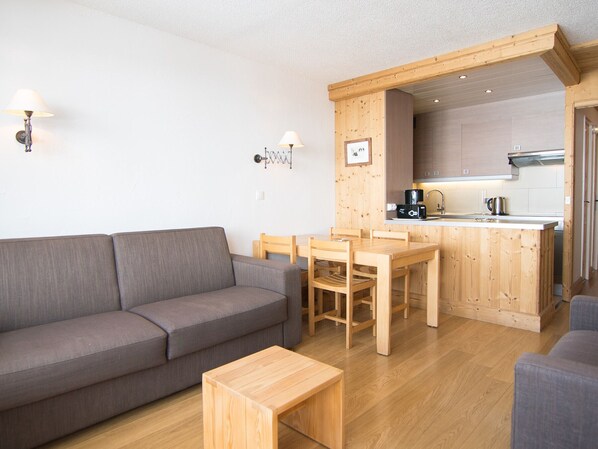Ein kompakter Wohnbereich mit grauem Sofa, Esstisch aus Holz und Stühlen, einem kleinen Holztisch und einer Küchenzeile mit Holzausbau und Spüle.