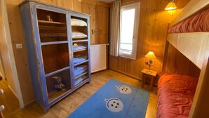 A room with wooden walls, a bunk bed, a wardrobe with glass doors, a window with curtains, and a small bedside table with a lamp. - Renovated 3-room apartment, south/west facing, near shops (Tignes)