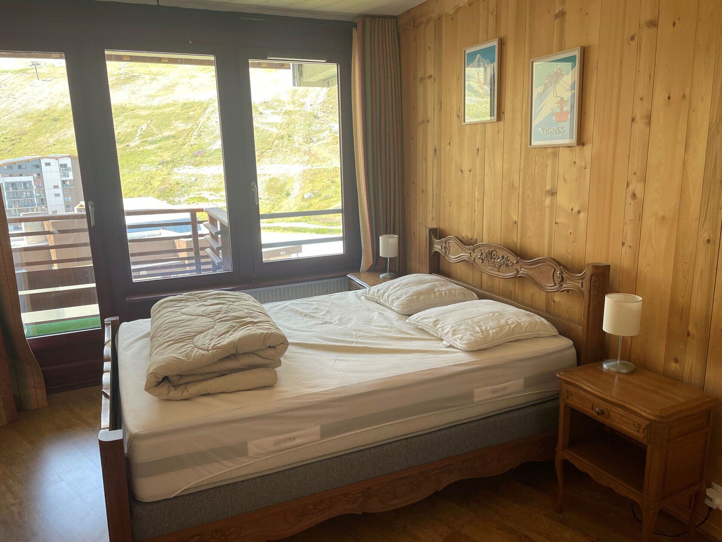 Une chambre à coucher avec une tête de lit en bois, un lit avec une literie blanche, deux lampes de chevet et une vue sur un balcon aménagé avec du mobilier d’extérieur.