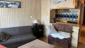 A cozy living area with a wooden panel wall, a sofa, a small table, and a kitchenette with blue-tiled backsplash. - Renovated 3-room apartment, south/west facing, near shops (Tignes)