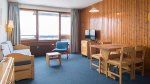 Huoneessa on puupanelilla verhoiltu seinä, televisio puukehikkoisessa kaapissa, sininen tuoli, puinen pöytä tuoleineen, sohva ja ikkuna, jossa on verhot.