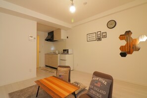 A modern living room with a wooden coffee table, two armchairs with American flags, a kitchen in the background, and a wall clock. - Modern Apartment Osaka Dome Mae (Osaka)