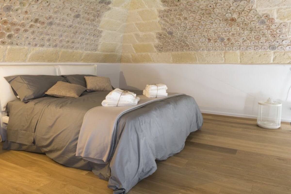 A neatly made bed with grey bedding, a stone wall with circular patterns, and a clear glass container on the floor.