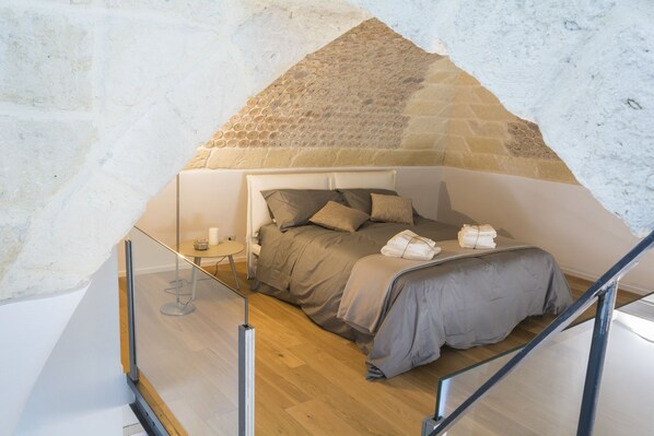 Un dormitorio con pared de piedra, una cama con ropa de cama gris, una mesita de noche con una vela y una barandilla de cristal.