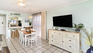 A dining area with a wooden table and white chairs, a kitchen with a fish-shaped sign, a TV on a cabinet, and a potted plant.