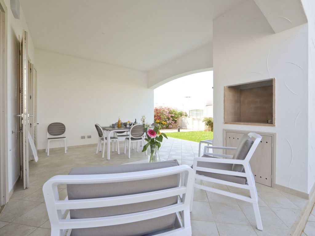 A covered outdoor seating area with white furniture, a grill, and a view of the garden.