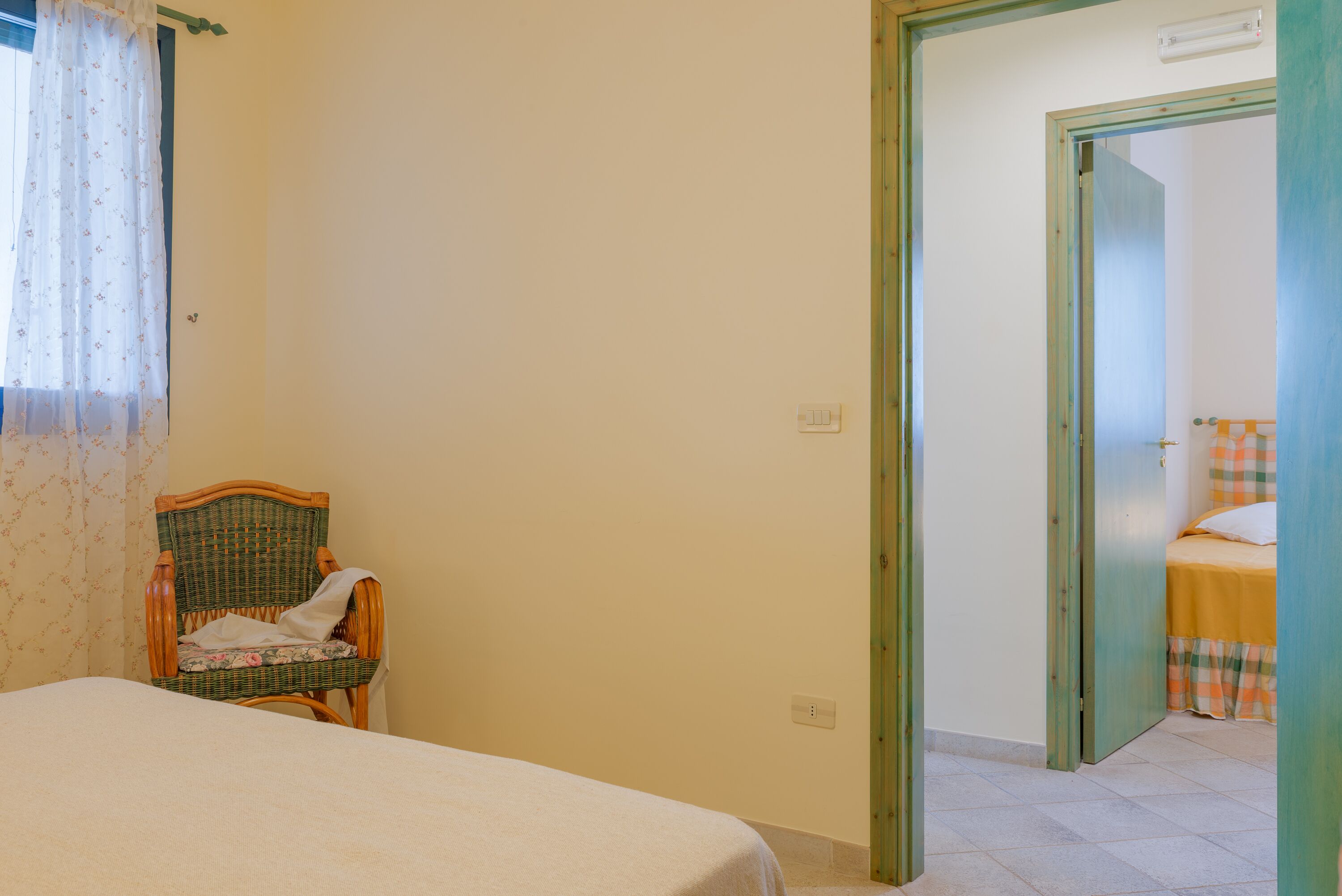 A bedroom with a bed, a wicker chair, and a door leading to another room.