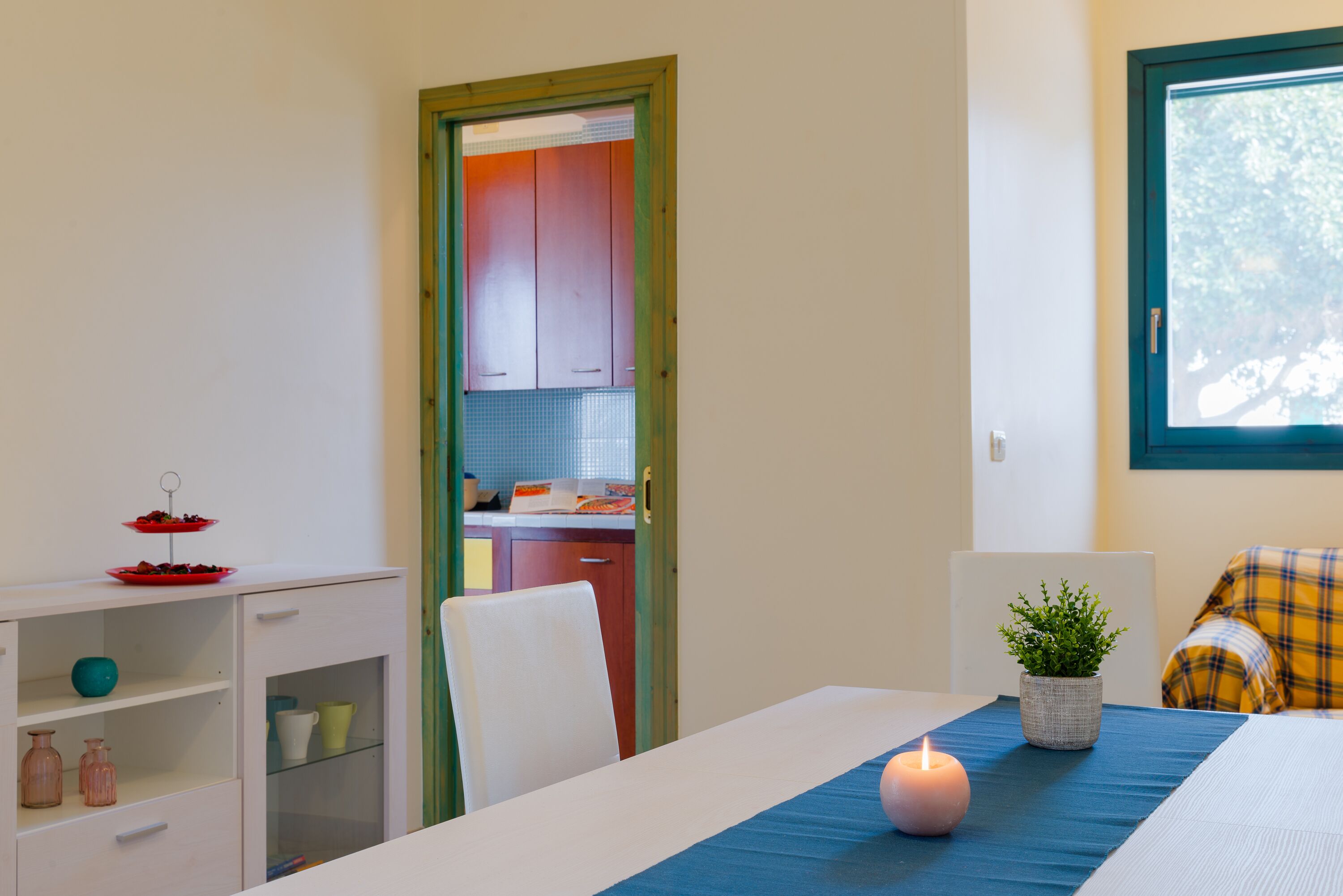 A room with a dining table, a candle, a potted plant, and a view of a kitchen through a door.