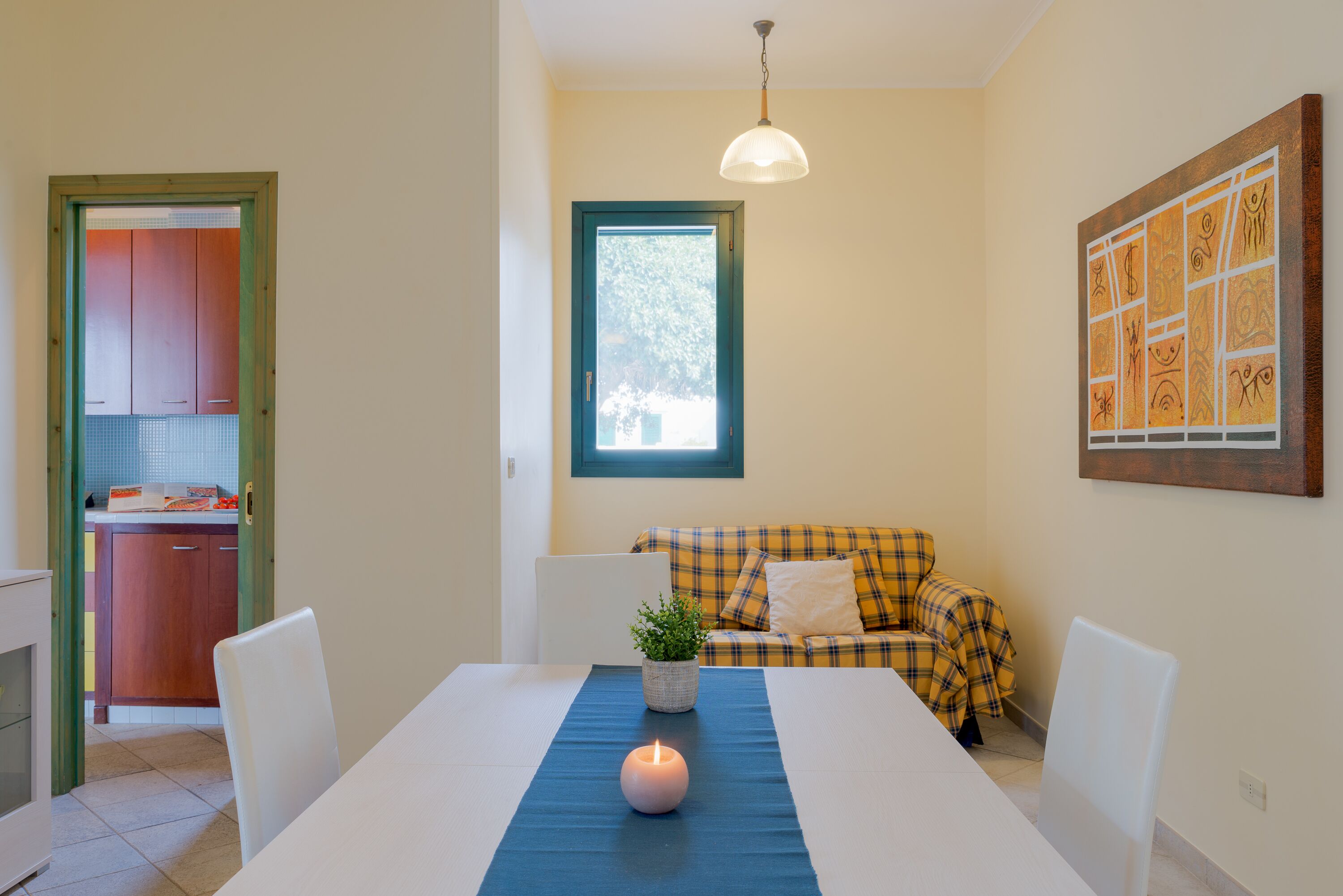 A dining area with a table, chairs, a candle, a plant, and a window with a view of greenery.