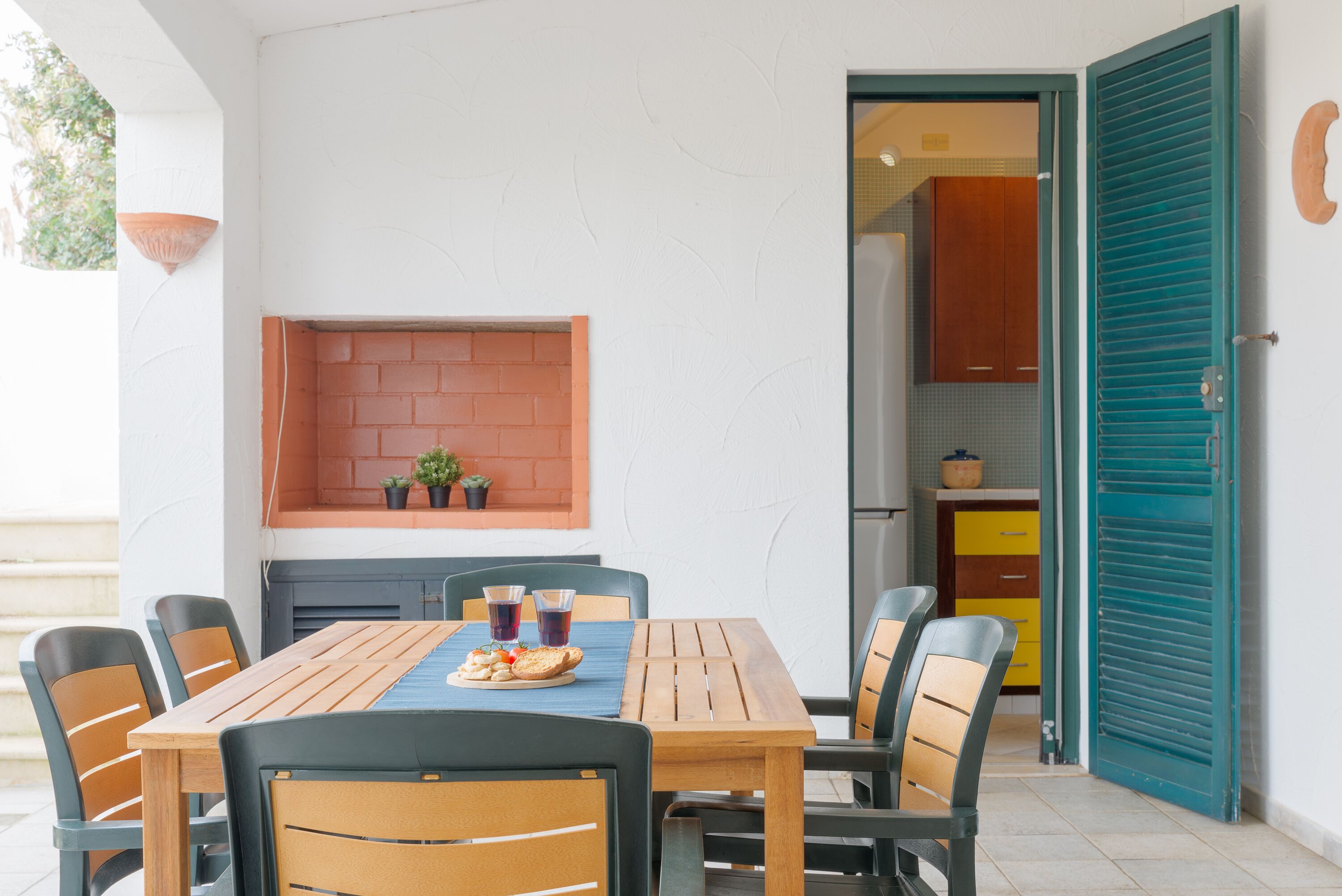 An outdoor dining area with a table and chairs, a brick shelf with plants, and a view into a kitchen with yellow cabinets and a teal door.