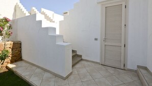 Een wit gebouw met een betegeld terras, een stenen muur en een deur met horizontale luiken.