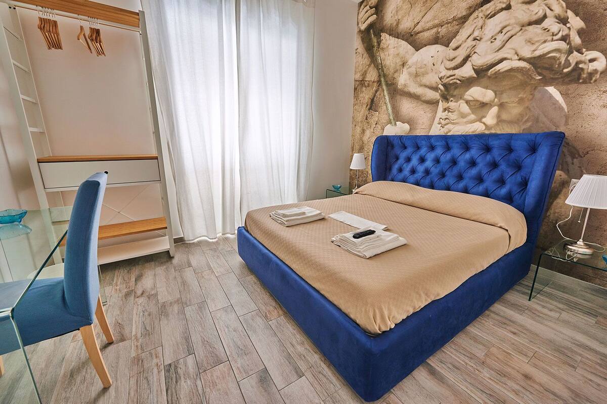 A bedroom with a blue upholstered bed, a wooden nightstand, a glass table, a chair, and a wardrobe with hanging clothes.
